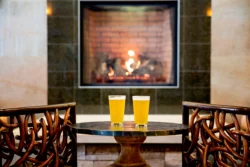 Two glasses of beer on a table between two chairs in front of a fireplace.