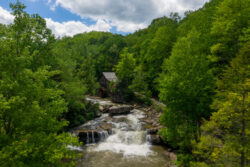 A scenic view of a watermill at Babcock State Park, surrounded by lush green trees. The mill sits beside a cascading waterfall under a partly cloudy sky, creating a peaceful and natural setting.