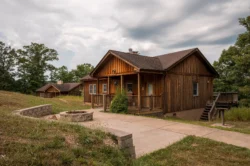 A rustic wooden cabin with a small porch sits on a grassy hill under a cloudy sky, reminiscent of the serene landscapes at Beech Fork State Park. A paved path leads to the front, and a fire pit is visible in the foreground. Another cabin is in the background surrounded by trees.