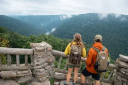Two hikers with backpacks overlooking forested canyon from stone overlook at Coopers Rock State Forest