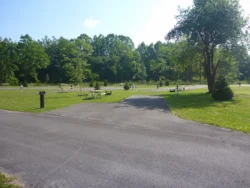 A paved road curves through a grassy campground with scattered trees, picnic areas, and fire rings on a sunny day.